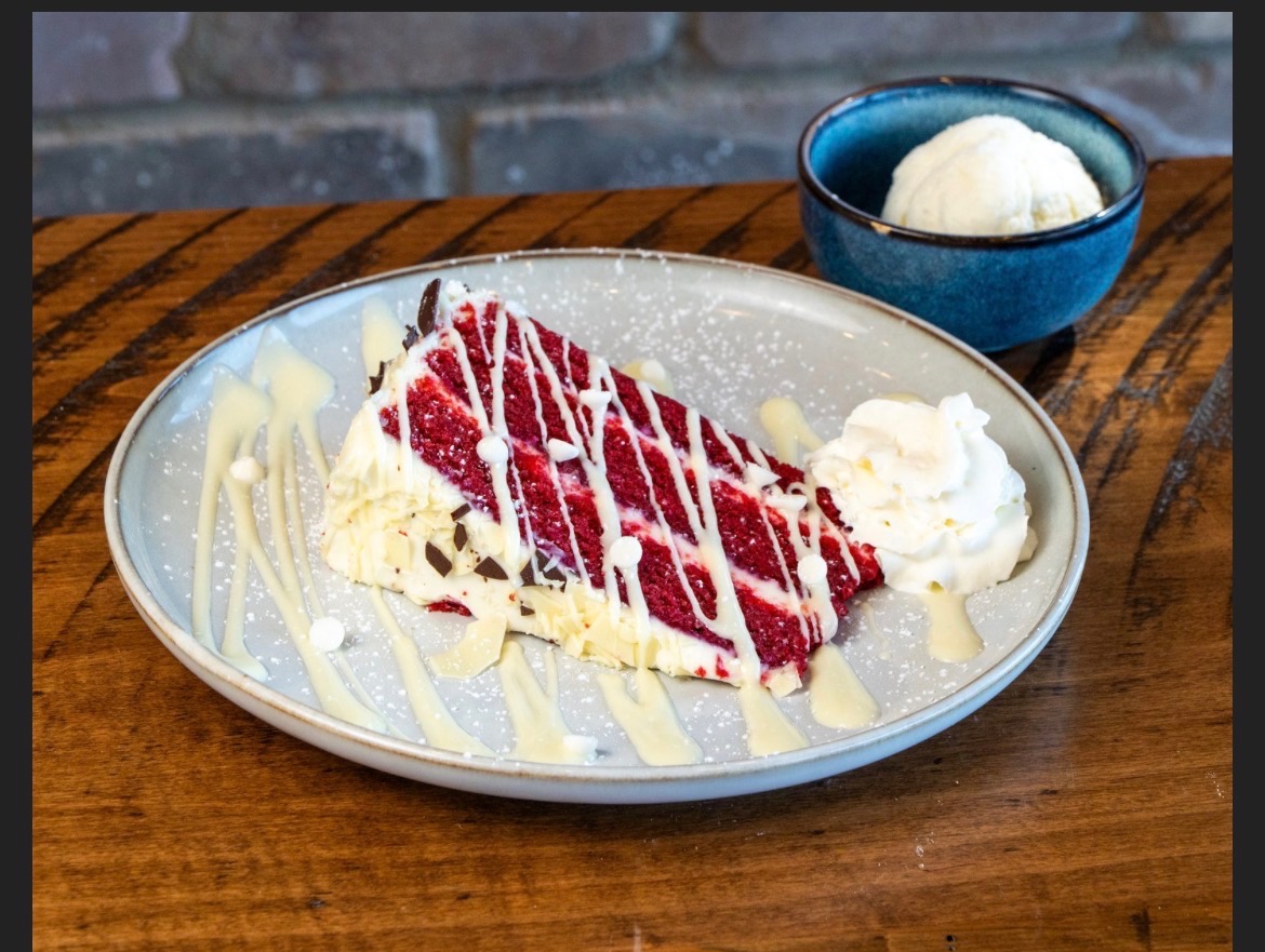 Slice of red velvet cake with white chocolate drizzle and a bowl of vanilla ice cream
